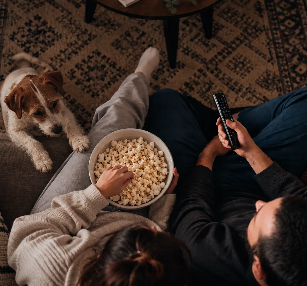 Una pareja ve una serie tumbada en el sofá, con palomitas y su perro